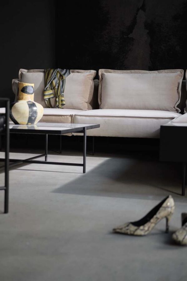 En moderne stue med en beige sofa med puder, et lille sort TomorrowsDesign sofabord, en stribet vase og et par højhælede sko på gulvet mod en mørkegrå kulisse.