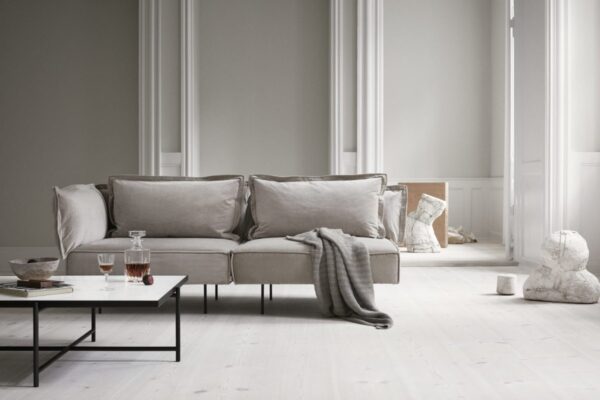 Elegant stue med en grå sofa, spredte hynder, et sort sofabord med glas og skål, et lysegrå gulvbelægning og hvidt gulv. Høje hvide søjler og solbeskinnede vinduer i baggrunden fremhæver TomorrowsDesign-æstetikken.