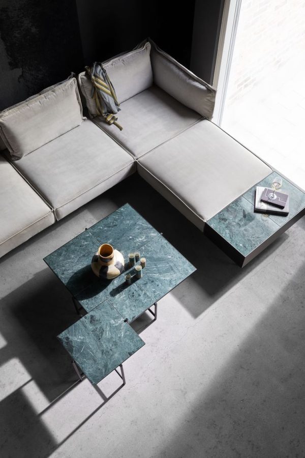Moderne stue med en hvid hjørnesofa draperet med en beige plade, to firkantede grønne marmor sofaborde, det ene med et tesæt og en bog. Sollys kaster en skygge gennem et stort vindue på betongulvet, hvilket viser TomorrowsDesigns æstetik.