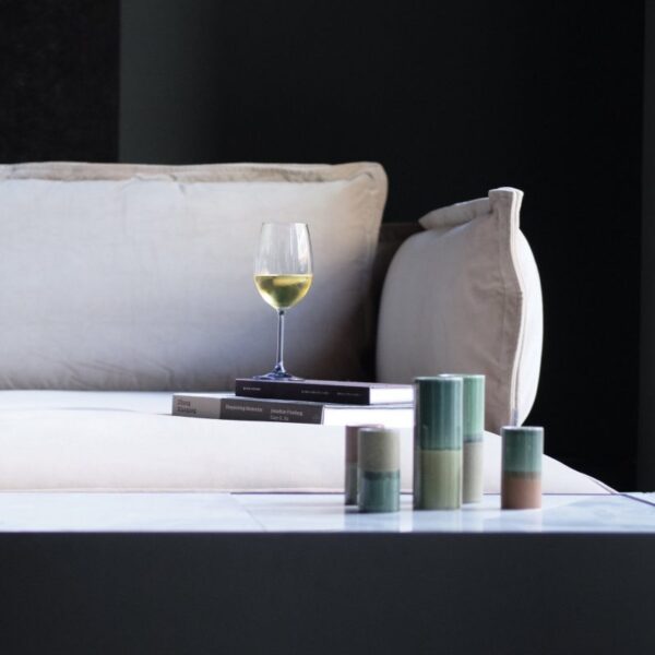 En moderne stuescene med et glas hvidvin ovenpå stablede bøger på et lavt bord ved siden af dekorative stearinlys. En plyssofa med en beige pude er delvist synlig i baggrunden, forstærket af elegante designmøbler.