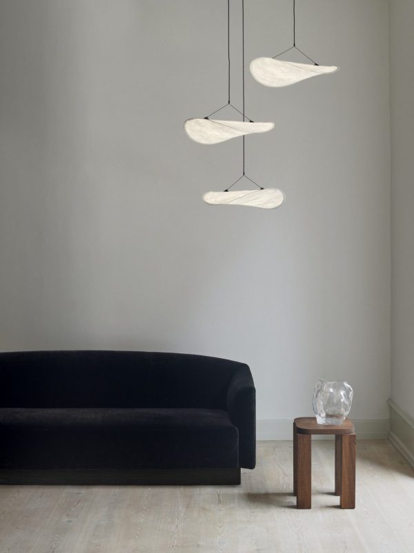 En minimalistisk stue med en mørk, buet sofa, et lille sidebord i træ og tre moderne NYHEDER - Tense Chandelier Ø55 hængelamper. Rummet har lyse grå vægge og trægulve.