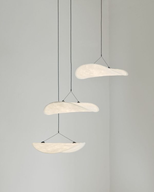 Tre NYE VÆRKER - Tense Chandelier Ø55 pendellamper ophængt i et loft med tynde kabler, mod en blød grå baggrund. lysene udsender et blidt, varmt skær.