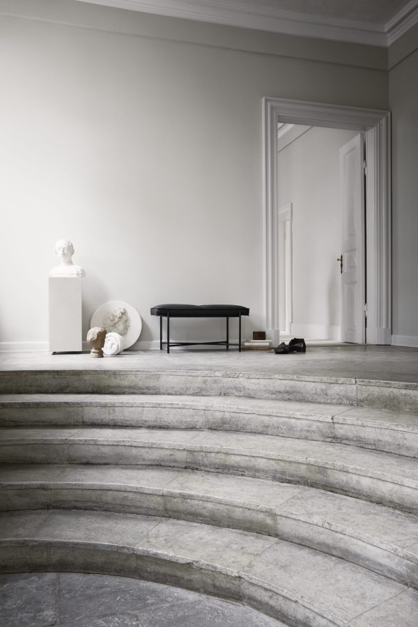 Et minimalistisk interiørdesign med en halvcirkelformet trappe med stentrappe, der fører til et elegant, sparsomt møbleret værelse med en sort bænk, klassiske skulpturer på sokler og en åben dør.
