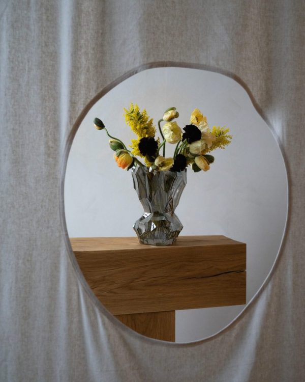 En buket blomster i et HEIN STUDIO - Refleksvase - Metallisk reflekteret i et rundt spejl, sat på et træbord mod en neutral stofbaggrund. blomsterne omfatter gule, sorte og hvide blomster.