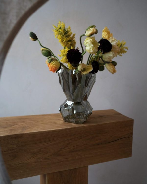 En buket tørrede blomster, inklusive sorte og gule blomster, arrangeret i et HEIN STUDIO - Refleksvase - Metallisk på et træbord, delvist reflekteret i et rundt spejl i baggrunden.