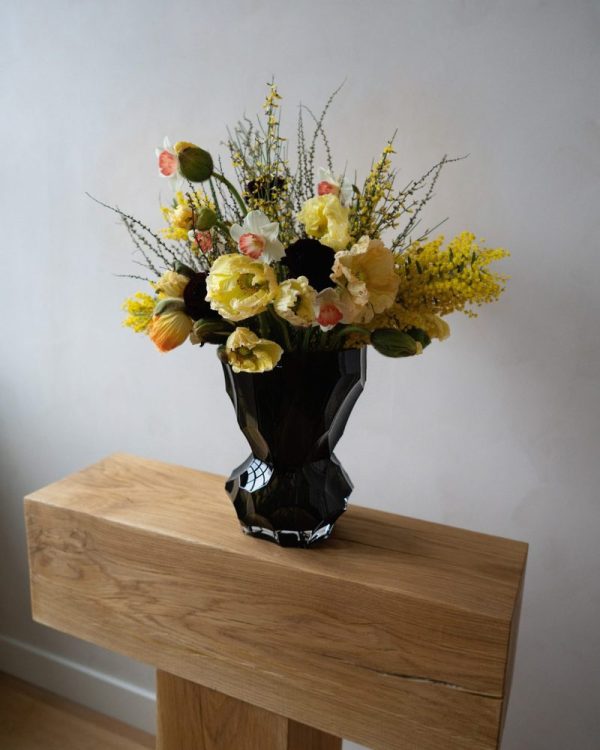 Et levende blomsterarrangement i et HEIN STUDIO - Reflection Vase - New Smoke, med gule og orange blomster med grønt, placeret på et lyst træskab mod en neutral væg.