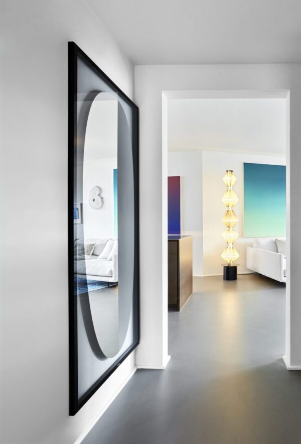 Et moderne, minimalistisk interiør i et hus med hvide vægge og et slankt sortrammet rundt spejl. Udsigten gennem en døråbning viser et rum med stilfulde designmøbler og farverige vægpaneler.