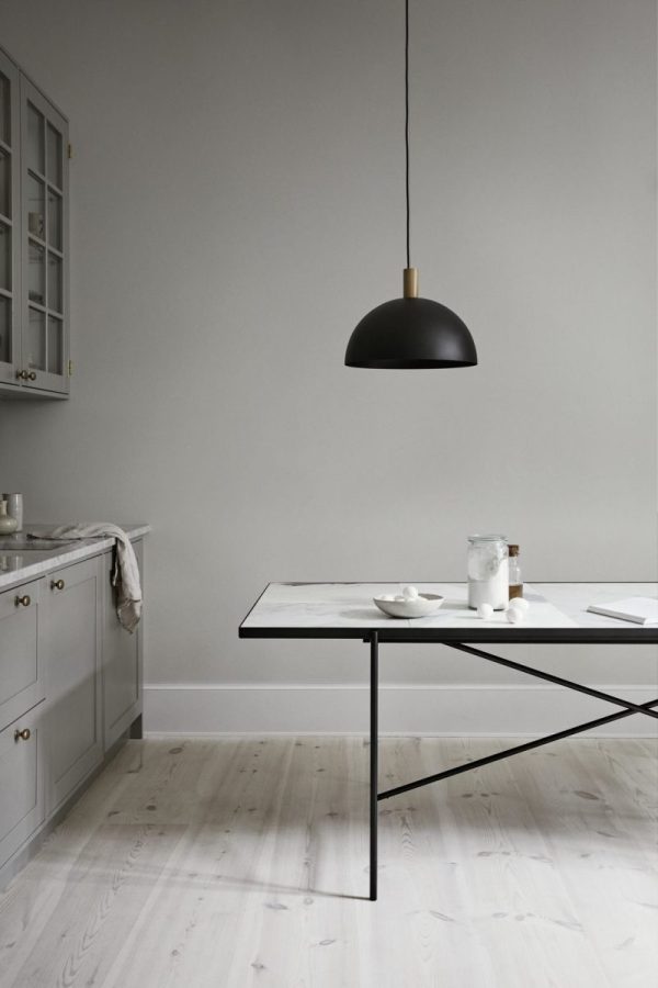 En minimalistisk spisestue med et elegant sort bord, hvide fade og en enkelt overhead sort pendel. Lyse trægulve og grå vægge komplementerer det enkle, moderne interiør.
