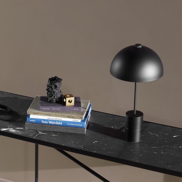 En moderne designbordslampe med en rund, sort skærm står på et marmorbord ved siden af en stak indbundne bøger. Et lille skulpturelt objekt og en dekorativ kube pryder også bordpladen.