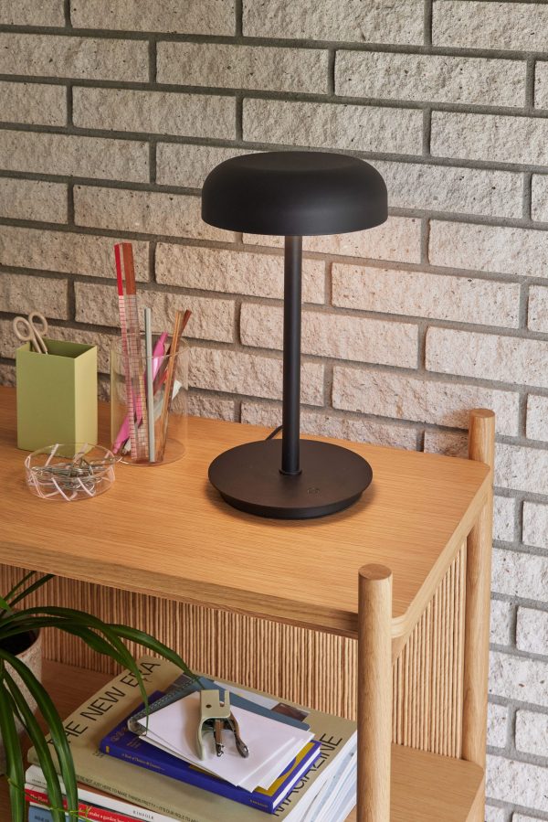 En moderne sort skrivebordslampe på en træhylde nær en murstensvæg. Hylden rummer også papirvarer, bøger og en lille potteplante fra TomorrowsDesign.