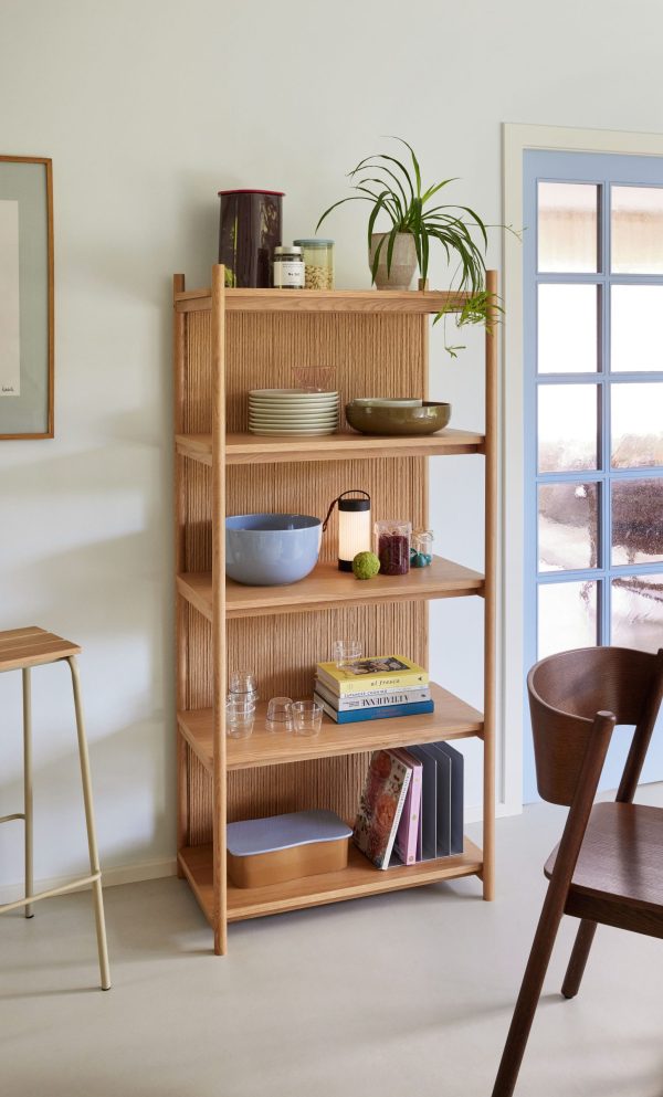 En fem-lags bogreol i træ i et minimalistisk rum, designet af TomorrowsDesign, fyldt med stueplanter, bøger, skåle og dekorative genstande nær et vindue med udsigt til det fri.