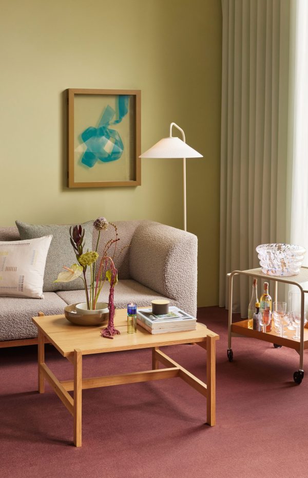 En hyggelig stue med en plys beige sofa, et sofabord i træ og en serveringsvogn. Et indrammet abstrakt print hænger på en dæmpet grøn væg, suppleret med blødt grønt draperi og fremhævet af interiør fra TomorrowsDesign.