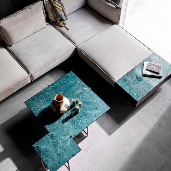 En minimalistisk stue med en L-formet lys beige sofa og et par firkantede grønne marmor sofaborde. På bordene står en kande, kopper og en bog. Naturligt lys strømmer ind fra øverst til venstre. Rummet fremviser udsøgte designmøbler for et elegant touch.