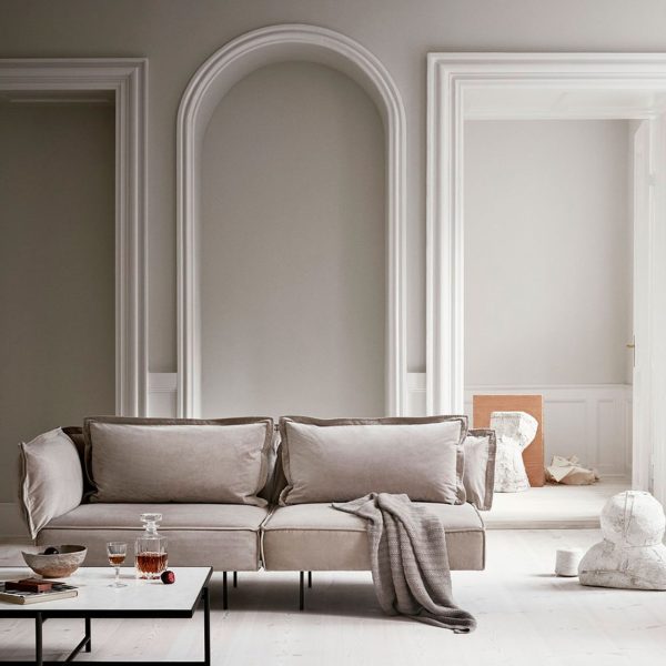 Elegant stue med en grå sofa med plyspuder, et lille sort sofabord med et glas og en bog og klassiske hvide vægge med dekorative buegange, stylet af TomorrowsDesign.