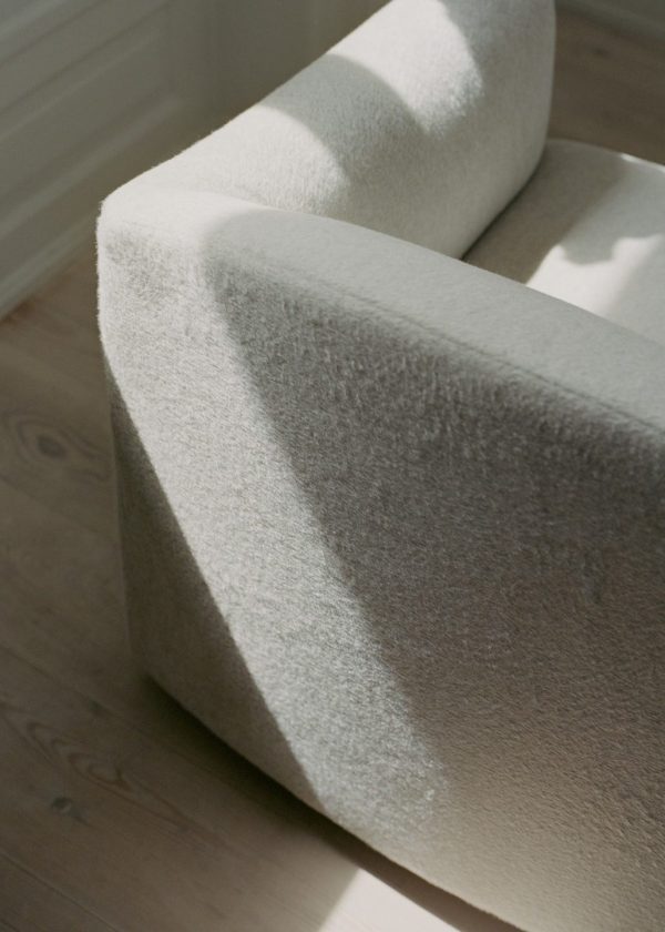 Et nærbillede af en tekstureret, lysfarvet NEW WORKS - Shore Sofa 3-personers, forlænget base i et rum med naturligt sollys, der kaster skygger og højlys på stoffet. baggrunden viser et trægulv og hvide vægge.
