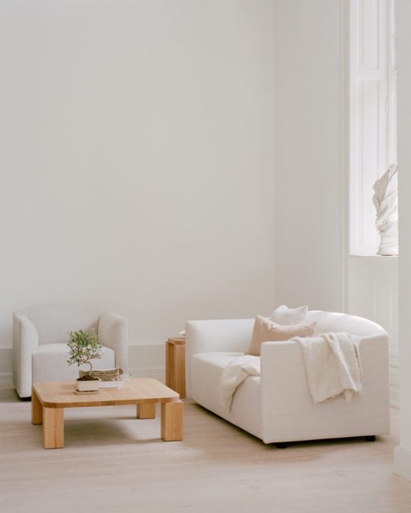 En minimalistisk stue med en neutral farvepalet med to hvide lænestole, NEW WORKS - Shore Sofa 2-personers, Extended Base, og et sofabord i træ med en lille plante på toppen, sat mod en baggrund af hvide vægge og et lyst træ. etage.