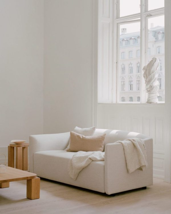 En minimalistisk stue med NEW WORKS - Shore Sofa 2-personers, Extended Base prydet med puder og et tæppe, et træsidebord og et stort vindue med udsigt over byens bygninger. En klassisk buste ses nær vinduet.