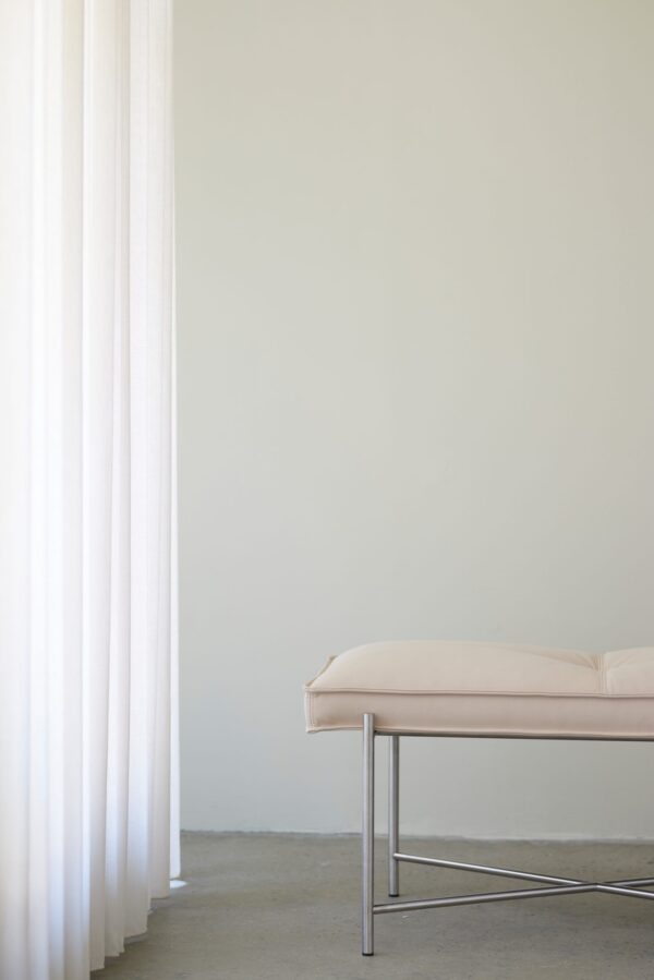 Et minimalistisk interiør med en lys pink bænk på et betongulv mod en almindelig hvid væg, delvist skjult af et rent hvidt gardin til venstre. Rummet omfavner elementer fra TomorrowsDesign og afspejler moderne interiør-æstetik.