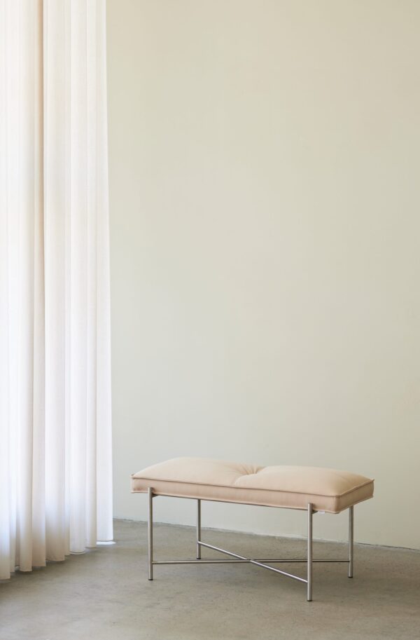 Et minimalistisk interiør med en beige skammel på en metalramme centreret foran et stort, lysfyldt vindue draperet med lange hvide gardiner, mod en bleggul væg.