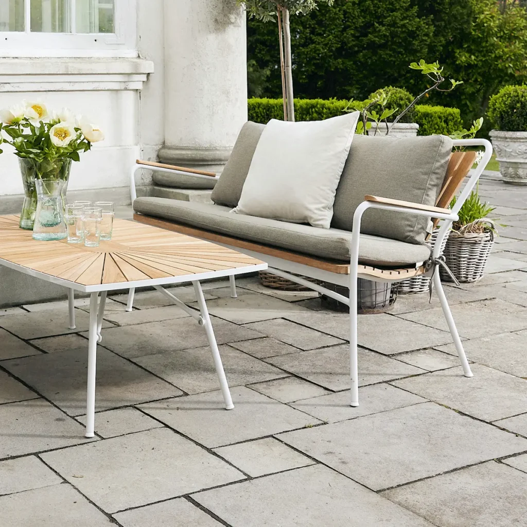Et udendørs opholdsområde med en moderne to-personers sofa med hynder, et rektangulært sofabord i metal og træ og en vævet kurv på en brolagt terrasse omgivet af frodige grønne områder.