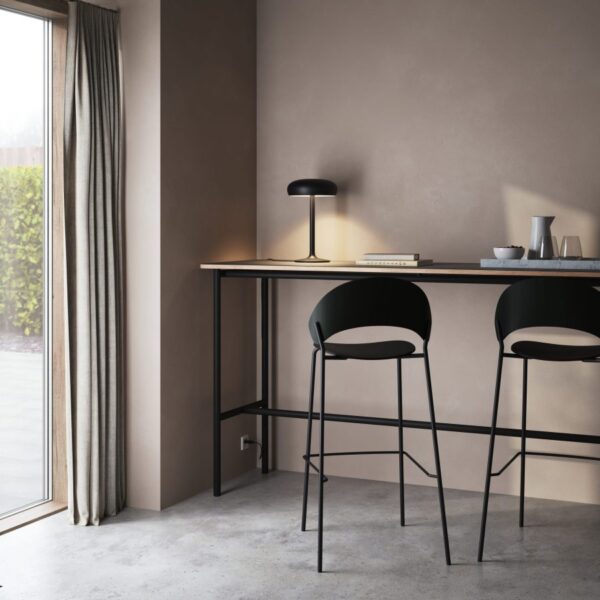 Et moderne hjemmekontor med et langt, slankt skrivebord og to EVA SOLO - Dosina Barstol H75 stole. der er en stilfuld lampe, nogle bøger og krus på skrivebordet, ved siden af et stort vindue med udsigt til en have og et draperet gardin på siden.