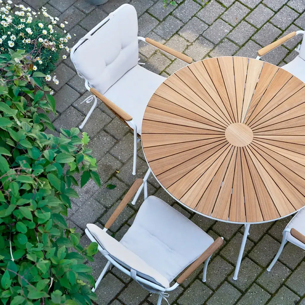 Luftfoto af et terrassesæt med et rundt træbord med radialt design og to hvide stole med metalstel, placeret på stenbelægning ved siden af frodige grønne buskadser.