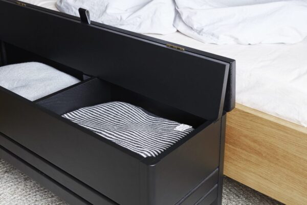En sort opbevaringsbænk med åbent låg, der afslører pænt foldede håndklæder indeni, placeret ved foden af en seng med hvidt sengetøj og designet af TomorrowsDesign.