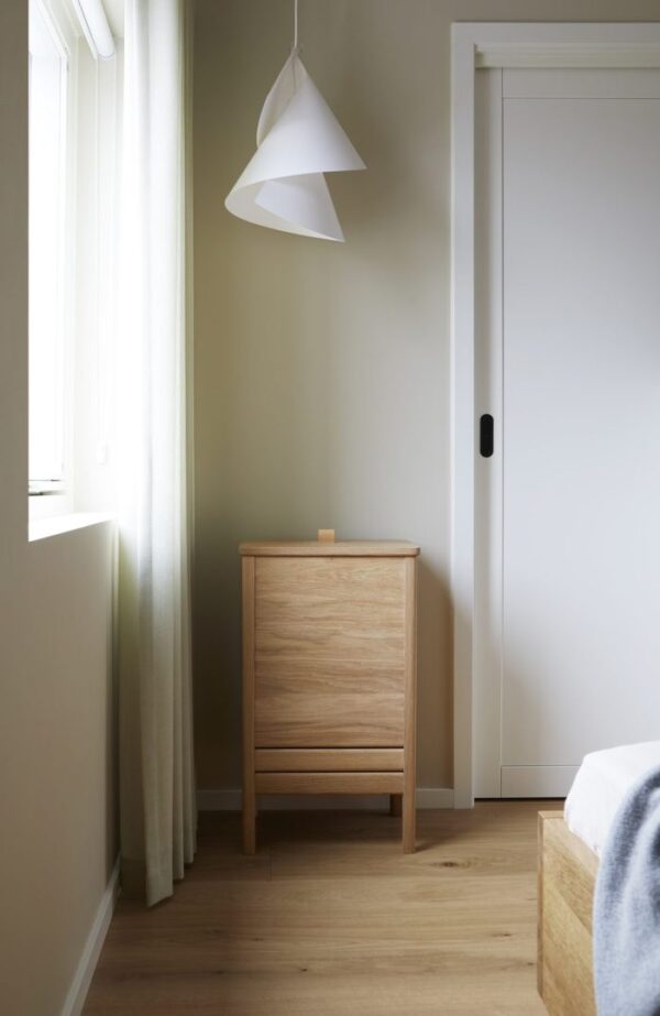 Et minimalistisk soveværelseshjørne med et lyst trænatbord ved siden af en seng med en hvid, kegleformet hængende lampe over hovedet. Et gennemsigtigt gardin dækker delvist et solbelyst vindue. Denne opsætning viser slank designmøbler-æstetik.