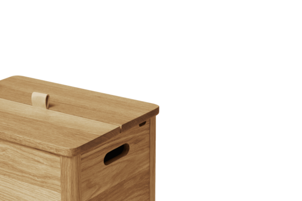 Nærbillede af en FORM & REFINE - A Line Vasketøjskurv med buet låg og cirkulært håndtag, sat mod sort baggrund. æsken har en glat finish og har et enkelt, rent design med en sideudskæring til at bære.
