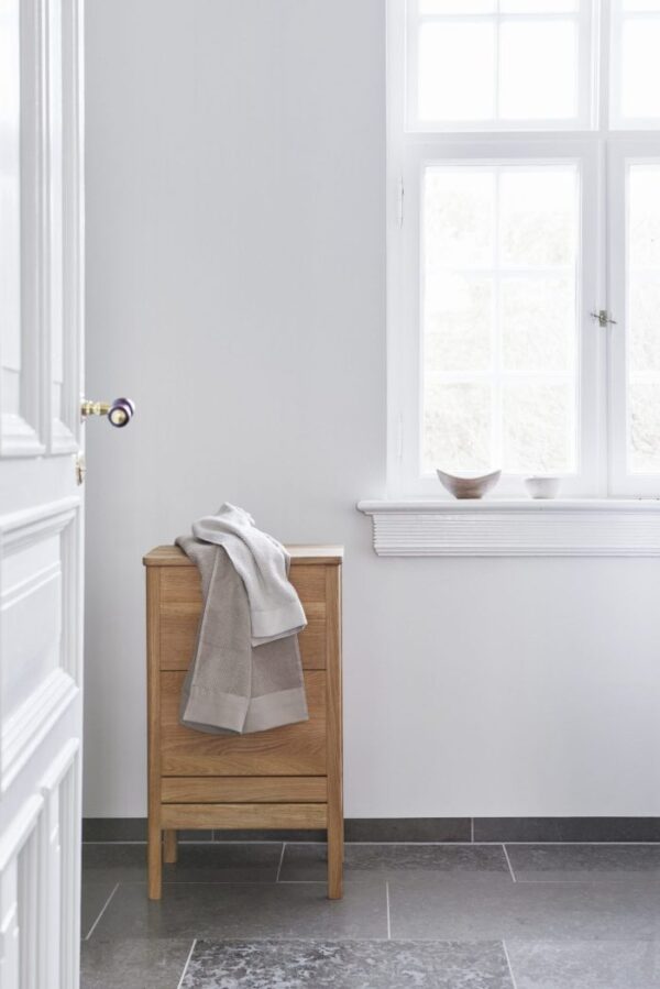 Et minimalistisk badeværelse med grå vægge og mørkt klinkegulv med FORM & REFINE - A Line Vasketøjskurv ved siden af et stort vindue med hvide rammer. et lyst håndklæde er afslappet draperet over Vasketøjskurv.