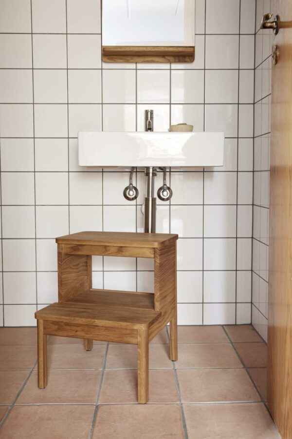 En trinskammel i træ foran en badeværelsesvask og et spejl, sat mod hvide flisevægge og interiør terracotta klinkegulv.