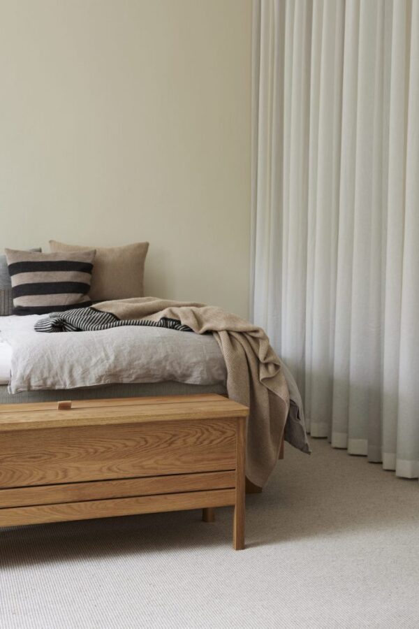 Et minimalistisk soveværelse med en træseng med sengetøj i neutrale toner, en FORM & REFINE - Aymara Plaid Stripes 190x130 pude og et draperet beige tæppe. rummet har lyse beige vægge og gulvlange gardiner.