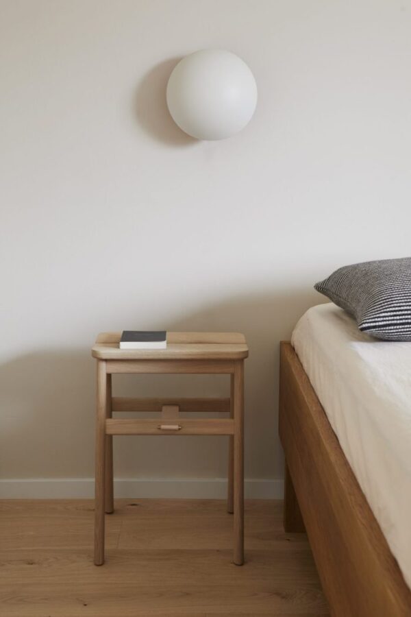 Et minimalistisk soveværelse med et natbord af træ med en skuffe og en bog på toppen, ved siden af en seng med en gråstribet pude. En sfærisk hvid væglampe er monteret over bordet, som eksemplificerer den elegante designmøbler-æstetik.