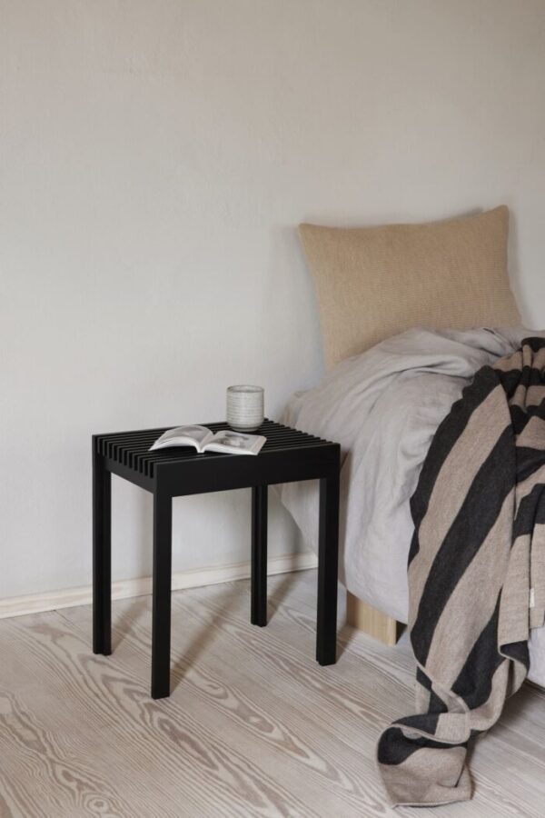Et minimalistisk soveværelseshjørne med en FORM & REFINE - Letvægts-taburet, der holder en åben bog og et stearinlys, ved siden af en seng med beige sengetøj og et sort-hvid-stribet plaid.
