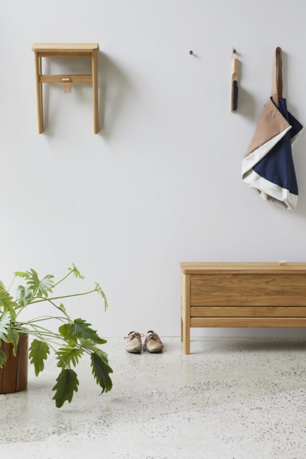 En minimalistisk entré med en hvid væg med en træbænk, en hængende skammel og værktøj på kroge er udtryk for TomorrowsDesign-æstetik. En stor potteplante står ved siden af bænken med et par sko på gulvet.