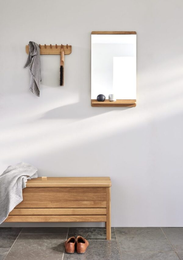 En minimalistisk entré med en træbænk, en FORM & REFINE - Rim Vægspejl på væggen og enkle kroge, der holder et håndklæde og børster. Et par sko er placeret pænt på gulvet, hvilket viser TomorrowsDesign indflydelse.