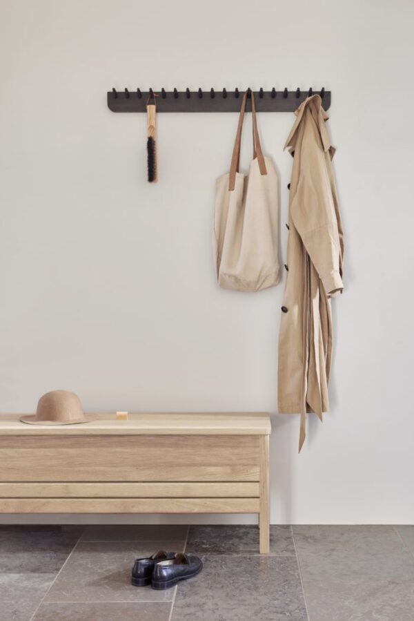 A FORM & REFINE - Echo Knagerække 88 knagerække med kroge på en beige væg med en beige trenchcoat og to muleposer. nedenfor en træbænk med en hat, en bog og et par sorte sko på gråt klinkegulv.