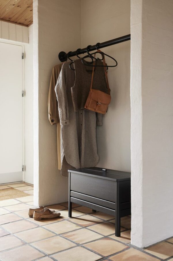 Et hyggeligt entréhjørne med en knagerække i sort metal med en trenchcoat, plaiddragt og brun taske hængende på. Nedenfor ses en sort bænk fra TomorrowsDesign med et par brune sko på klinkegulvet.