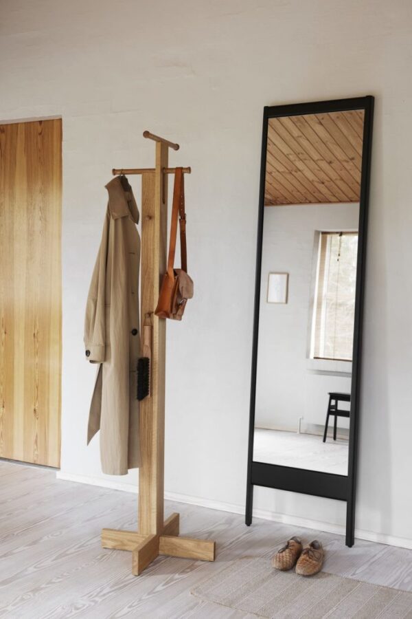 En minimalistisk entré med en FORM & REFINE - Foyer Stumtjener med en beige frakke og en lædertaske, et spejl med sort ramme i fuld længde og spredte sko på et lyst trægulv. blød naturlig belysning og et synligt træloft forstærker den fredfyldte atmosfære.