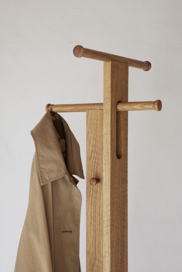 FORM & REFINE - Foyer Stumtjener med et minimalistisk design og en tan trenchcoat hængende på den ene arm, vist mod en almindelig hvid væg.
