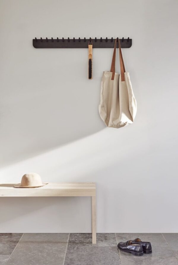 En minimalistisk entré med en FORM & REFINE - Lightweight Bænk, en hat på, en knagerække med en taske og en paraply og et par sko på et klinkegulv, oplyst af naturligt lys.