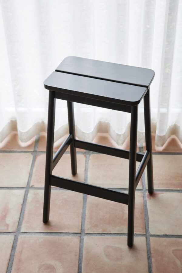 En enkel, sort FORM & REFINE - Angle Standard Barstool 75 står foran et hvidt gardin ved et klinkegulv, hvilket giver en kontrast til dets minimalistiske design og neutrale omgivelser.