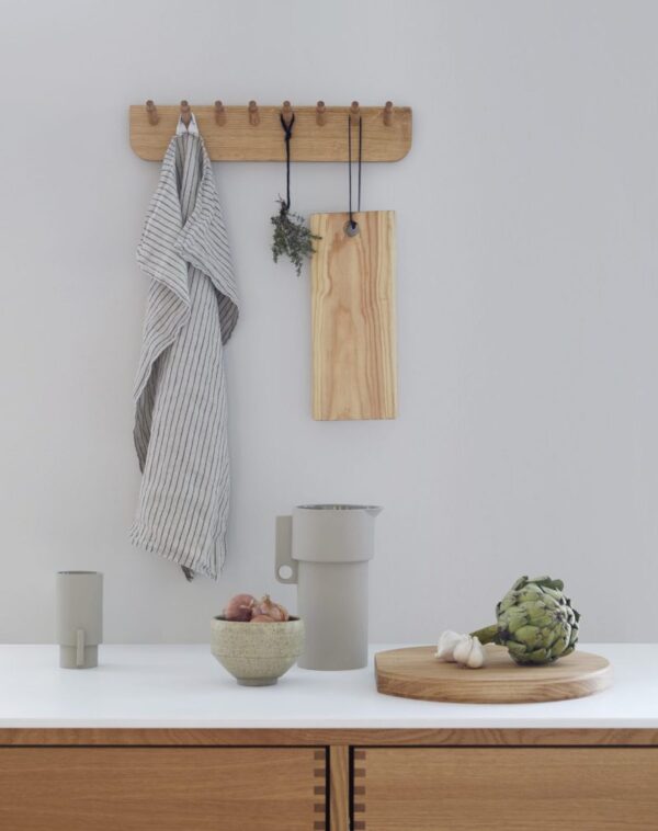 En minimalistisk køkkenscene med FORM & REFINE - Echo Knagerække 40 med et stribet håndklæde og en træplade, en bordplade med en keramisk kande, en skål med hvidløg og skalotteløg og en træbakke med en artiskok.