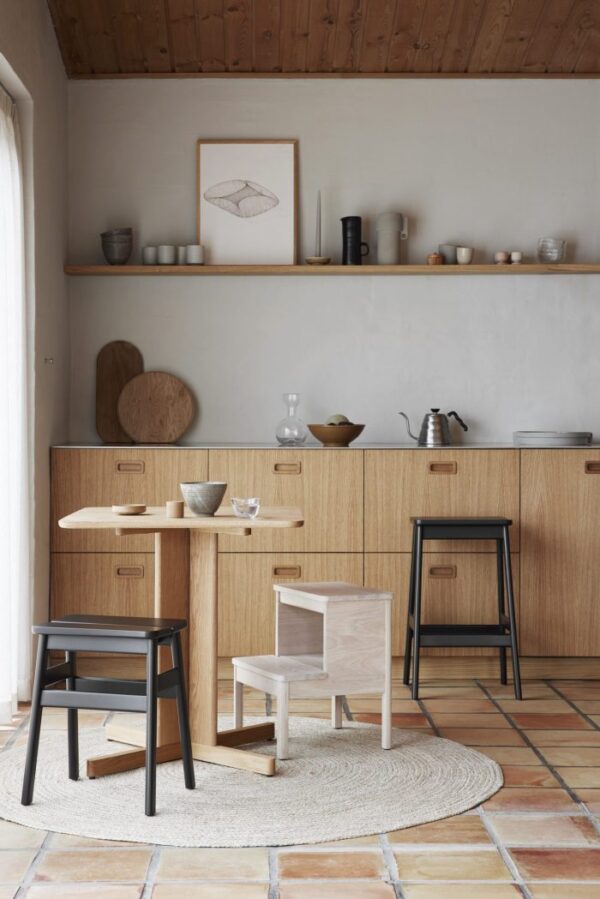 Et minimalistisk køkken med træskabe og hylder, med et udvalg af keramisk service. Der er et træbord med stole og en skammel på et rundt vævet tæppe over terracotta flisegulv. Dette interiør inkorporerer praktisk design, samtidig med at det bevarer en æstetisk appel.