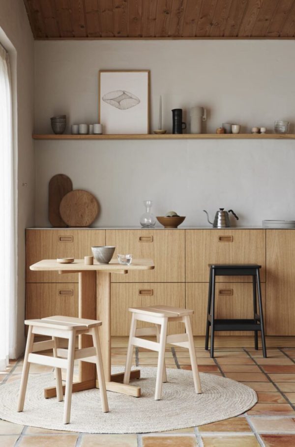 Et minimalistisk køkkeninteriør med en FORM & REFINE - Quatrefoil Bord 68x68 træbord med integreret opbevaring, to træskamler sat på et cirkulært tæppe og en sort skammel. fritlagte træbjælker i loftet og keramiske dekorationer fremhæver indretningen.
