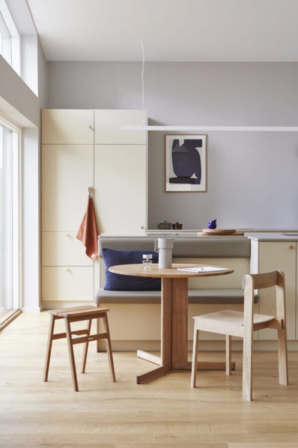 Et moderne køkken med lyse trægulve og crememøbler inkluderer en FORM & REFINE - Trefoil Bord Ø75, indbygget siddekrog med en blå polstret bænk, et træbord og en stol. Et minimalistisk maleri hænger på væggen.