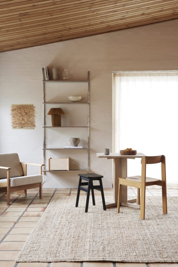 En minimalistisk stue med en naturlig farvepalet med et spisebord i træ, stole, en sort skammel og en reol mod en hvid murstensvæg. FORM & REFINE - Quatrefoil Bord 68x68 dækker gulvarealet.