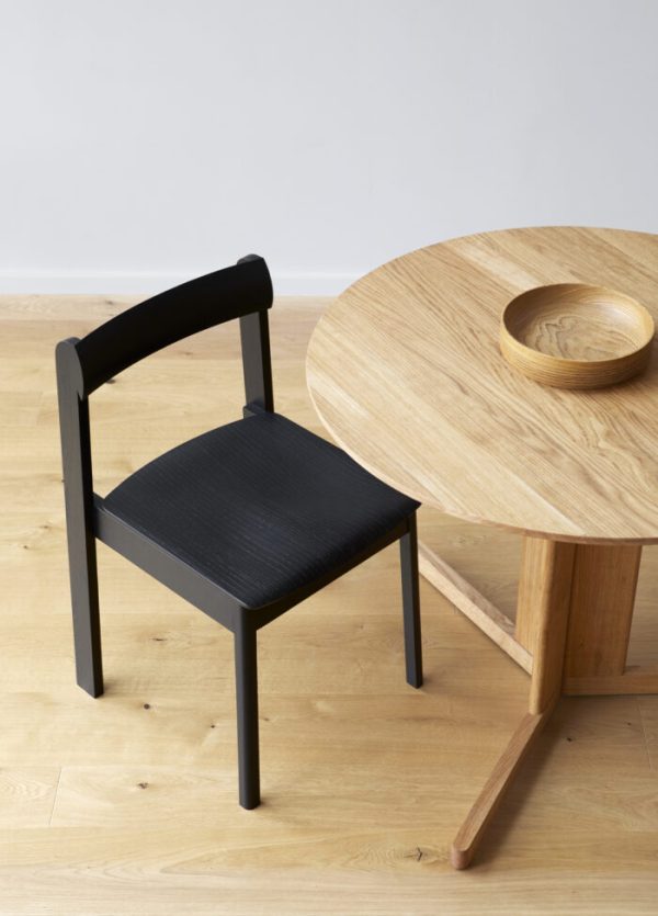 En minimalistisk indstilling med en FORM & REFINE - Trefoil Bord Ø75 med et tekstureret sæde ved siden af et rundt træbord på et lyst trægulv. En træskål hviler på bordet.