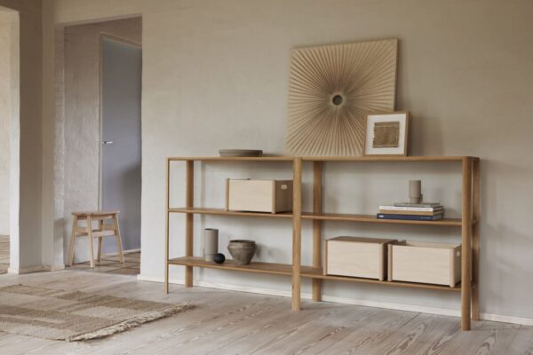 En minimalistisk stue med en FORM & REFINE - Leaf Reol 2x3 med kasser og indretning, en lille træskammel og et stort gulvspejl mod en tekstureret beige væg. Et vævet tæppe ligger på det lyse trægulv.