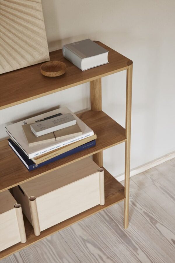 A FORM & REFINE - Leaf Reol 1x3 med to hylder, med pænt arrangerede bøger, en vævet kurv og en lille rund børste. bordet står op ad en almindelig væg med lyst trægulv.
